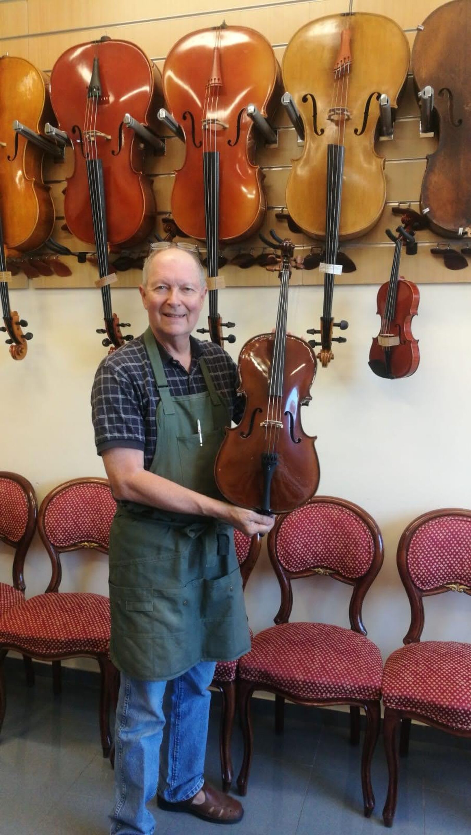 Dennis Braun in his atelier, holding a freshly varnished cello in front of a wall of instruments